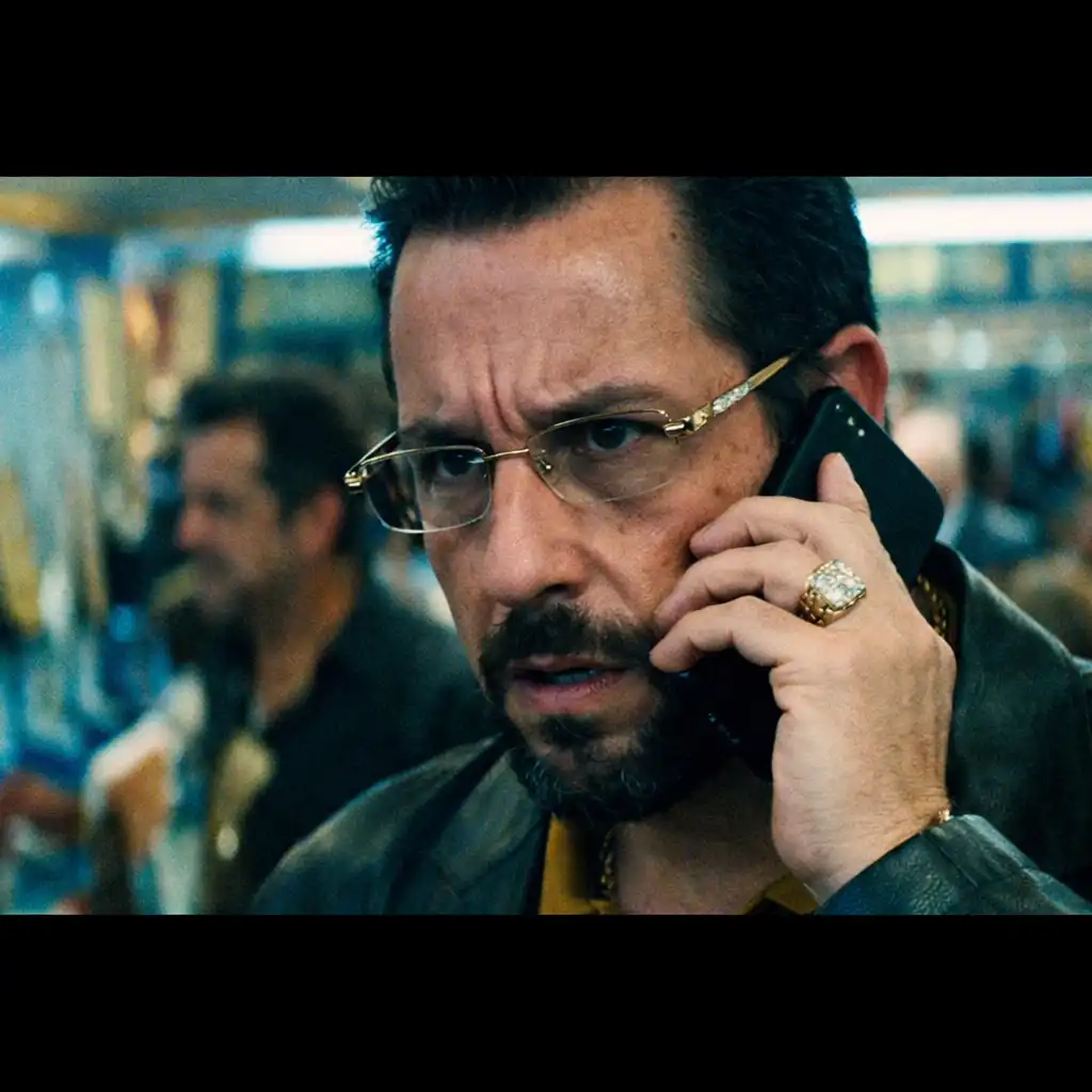 A still from Uncut Gems (2019) showing a man in a crowded jewelry store, his face tense, holding a phone to his ear amid cluttered display cases.